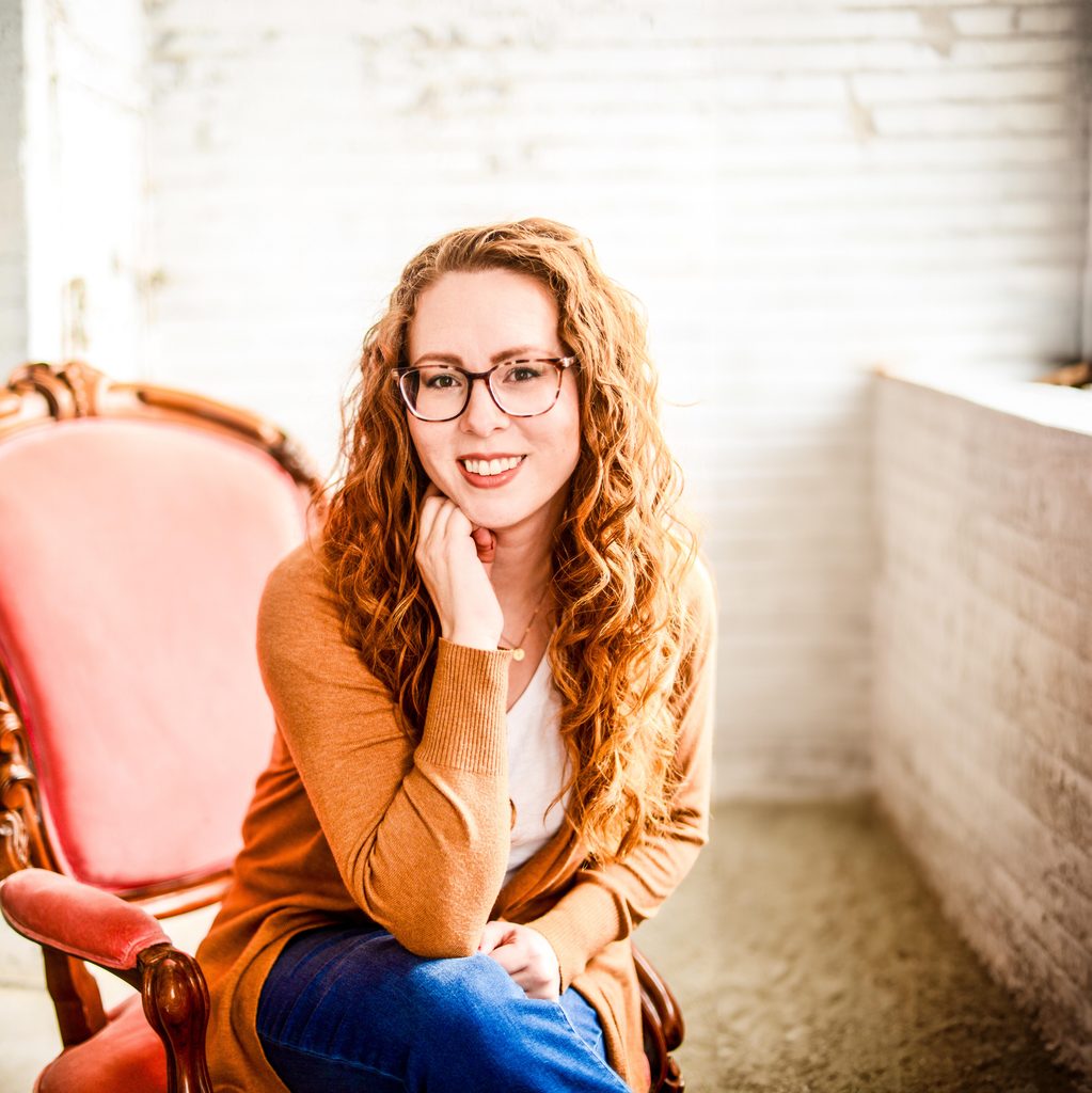 Headshot of brand designer with curly red hair sitting in a pink chair