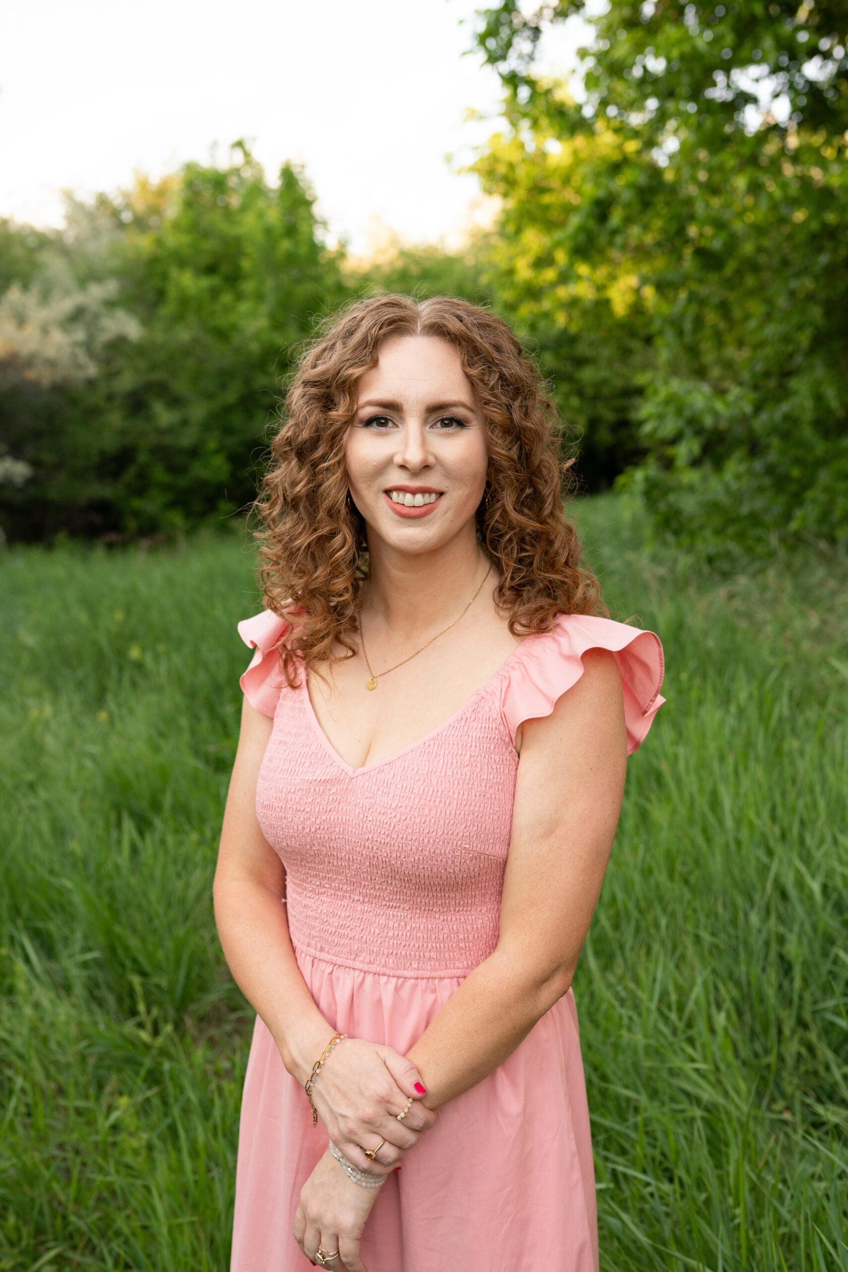 Graphic designer headshot, woman with curly red hair wearing pink dress with outdoor background
